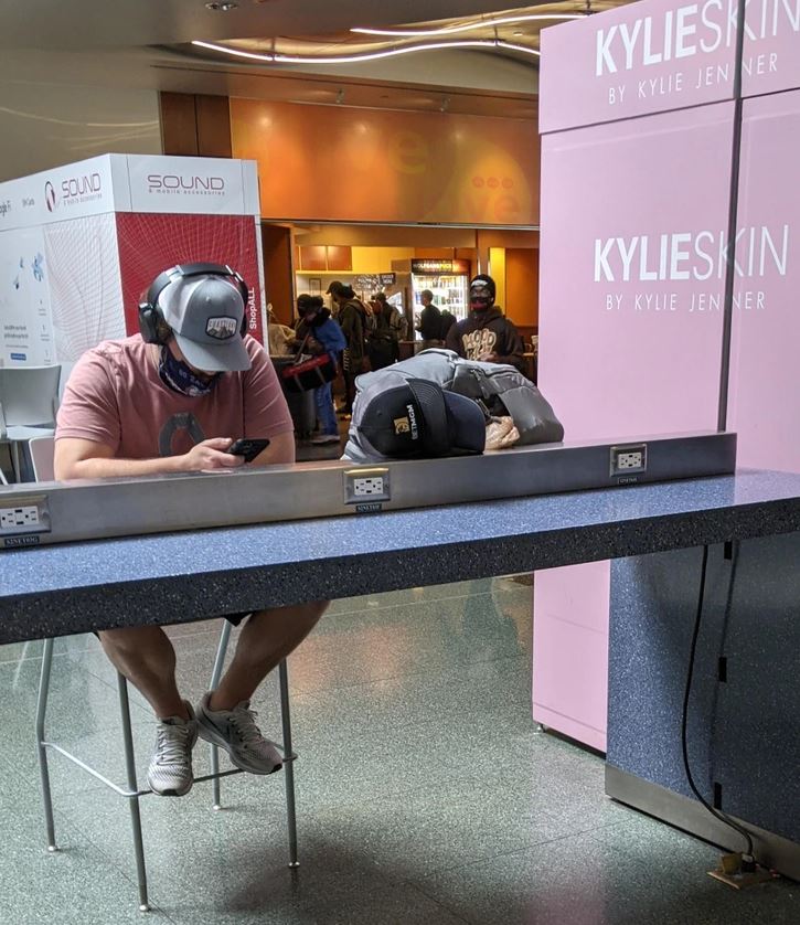 Muž s čiapkou a slúchadlami sedí pri nabíjacej stanici, zahĺbený do svojho telefónu na letisku.