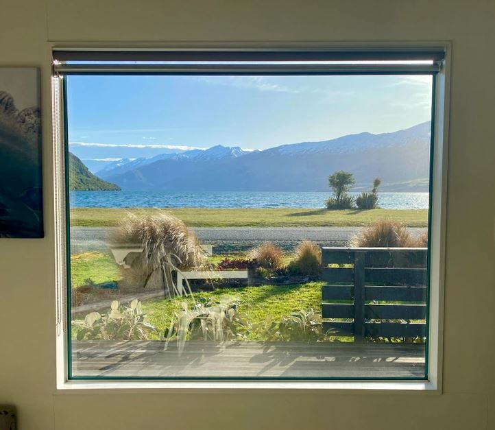 Pohľad z okna na pokojné jazero a horské vrcholy pod modrou oblohou.