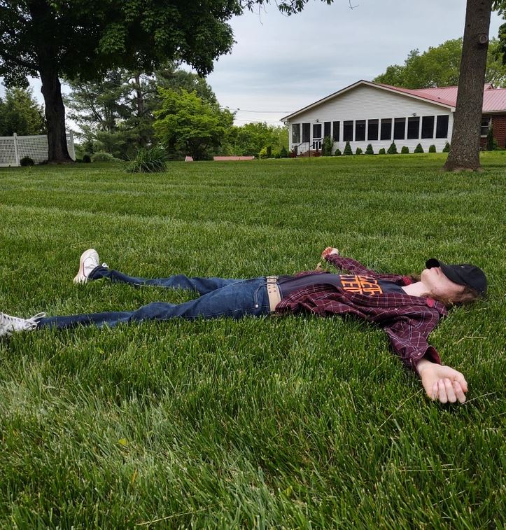 Osoba leží na tráve v záhrade pred bielym domom; uvoľnený oddych v prírode.