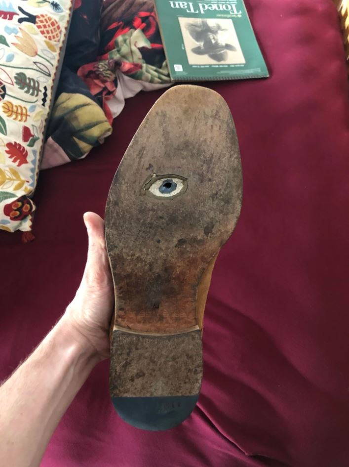 Podrážka topánky s kresbou oka, držiaca ruka nad červenou pohovkou s dekou a knihou.