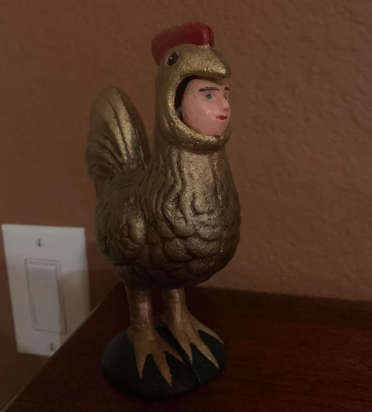 Soška kura s ľudskou hlavou, stojaca na drevenom povrchu pri svetelnom vypínači.