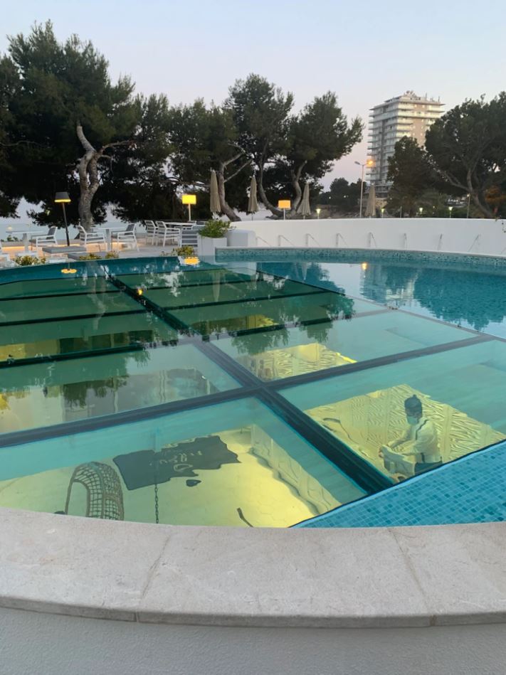 Odkrytý bazén so sklenenou podlahou nad podvodnou miestnosťou, vonkajšia terasa a stromy na pozadí.