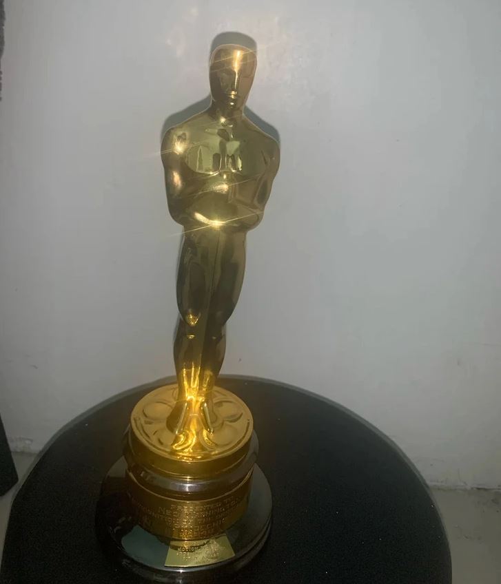 Zlatá socha na podstavci, pripomínajúca filmovú cenu, osvetlená, umiestnená na tmavom povrchu proti bielemu pozadiu.