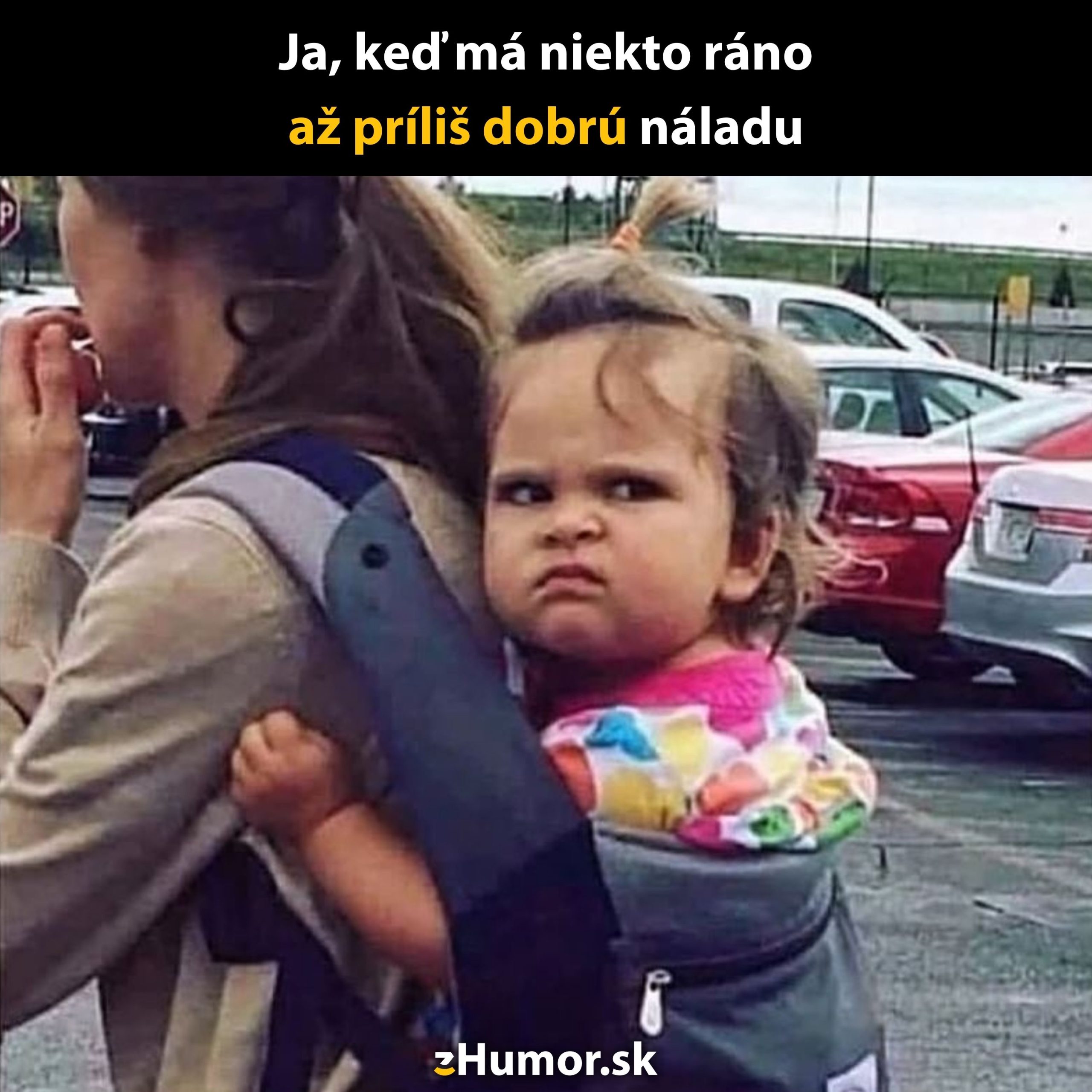 Dojča s nespokojným pohľadom v nosiči na chrbte matky na parkovisku.