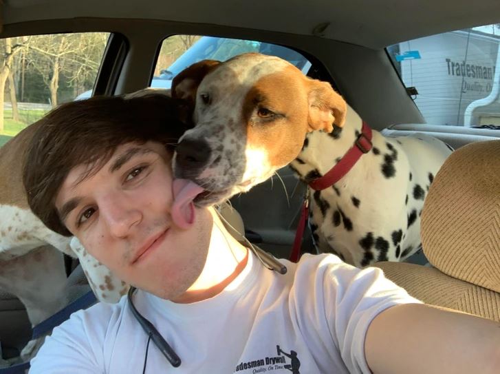 Muž v aute s úsmevom dostáva olíznutie od psa s fľakmi na bielej srsti, ktorý sedí vedľa neho.