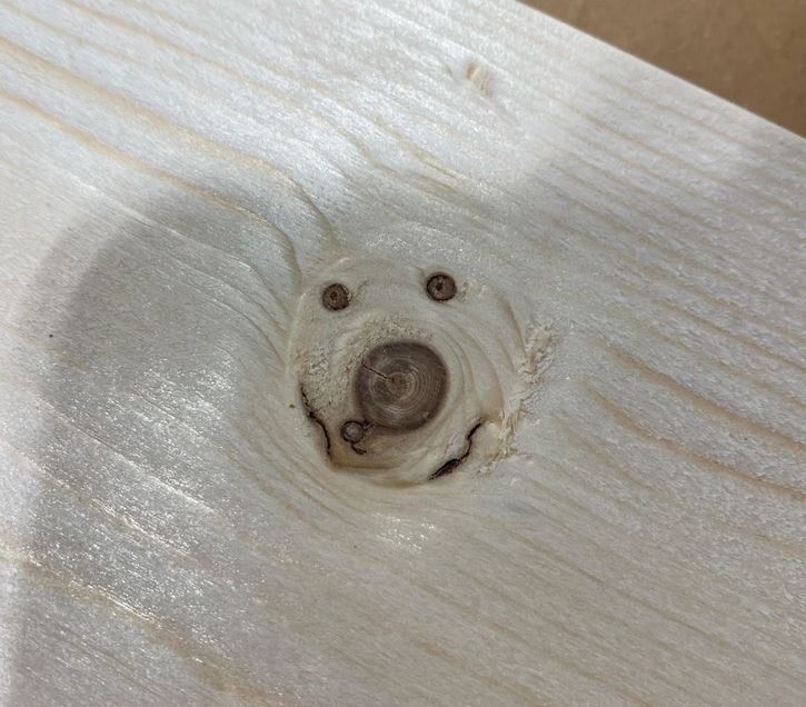 Drevený uzol na doske pripomínajúci úsmev, zblízka zachytávajúci textúru a detaily dreva.