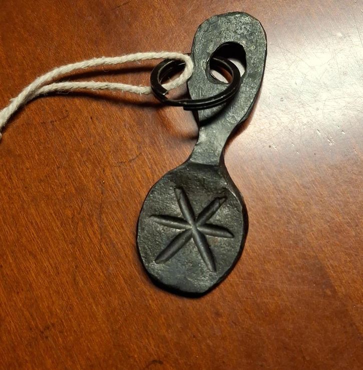 Kovový amulet s vyrytým symbolom na drevenom pozadí, uchytený šnúrkou.