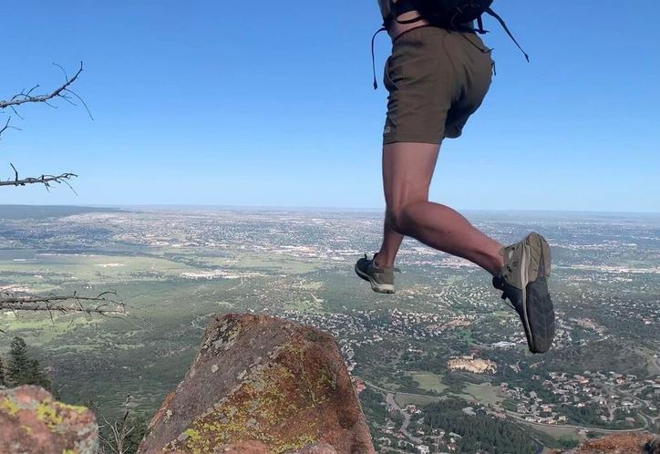 Skokan v horách: Muž vyskakuje z útesu s panoramatickým výhľadom na krajinu za ním.