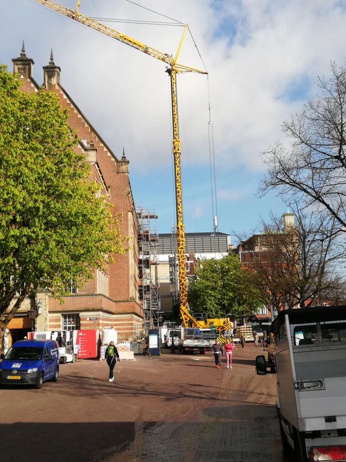 Žeriav pracujúci na stavbe pri historickej budove v meste, obklopený chodcami a stromami pod modrou oblohou.