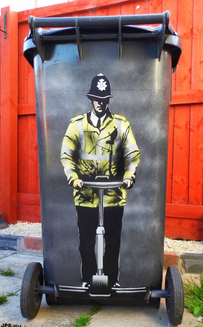 Grafiti policajta na segwayi na smetnej nádobe pred červeným plotom.