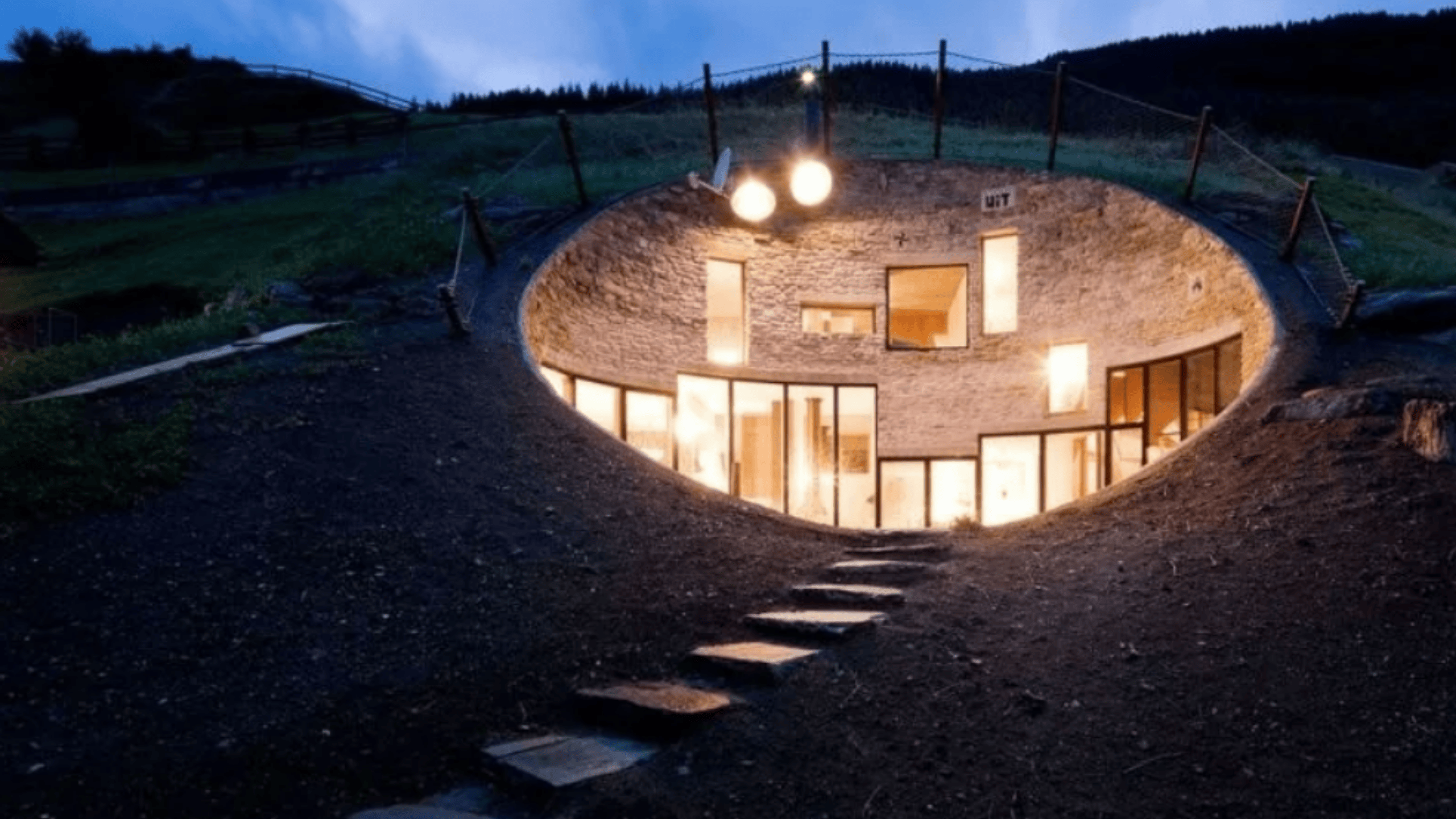 Moderný dom v zemi s osvetlenými oknami, zabudovaný do svahu v prírode pri západe slnka.