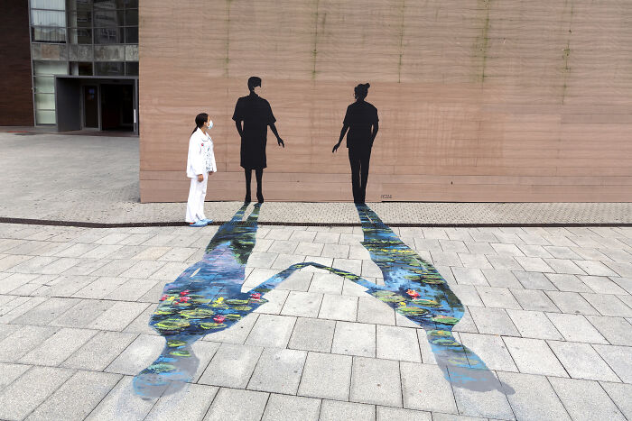 Žena v bielom plášti stojí pred múrom s nástennou maľbou a farebným tieňom na zemi.