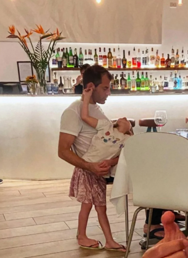 Otec v reštaurácii drží spiace dieťa, v pozadí barová polica s fľašami a kvety.