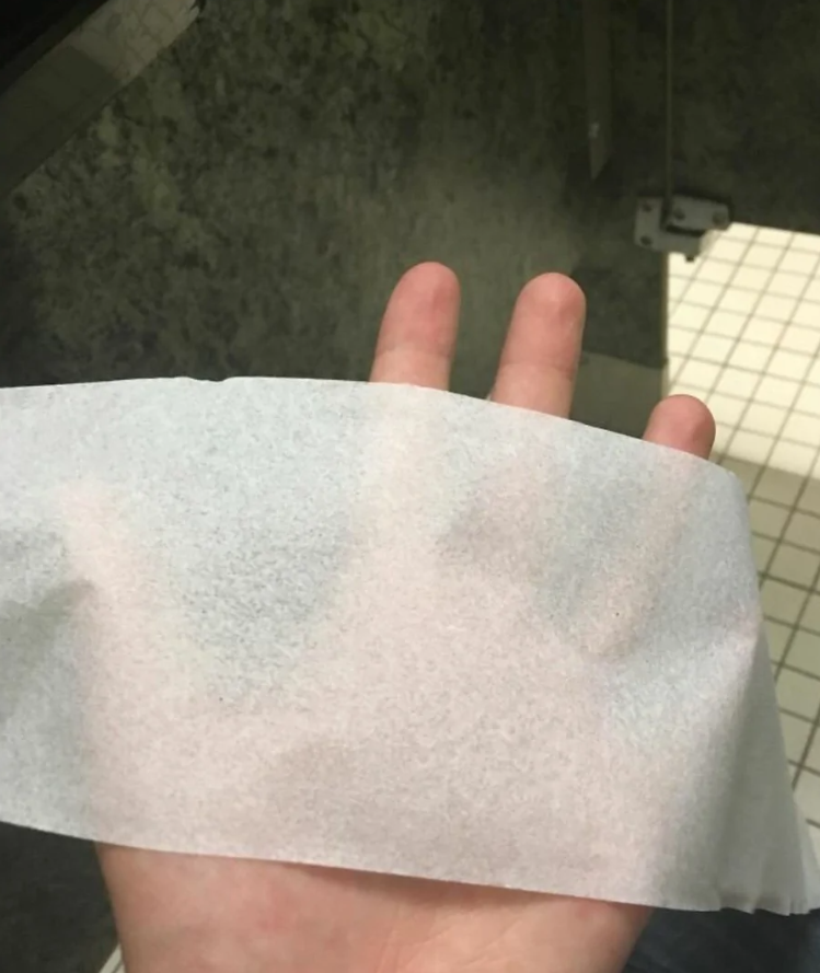 Ruka držiaca tenký útržok toaletného papiera na podlahe verejných toaliet s bielymi dlaždicami.