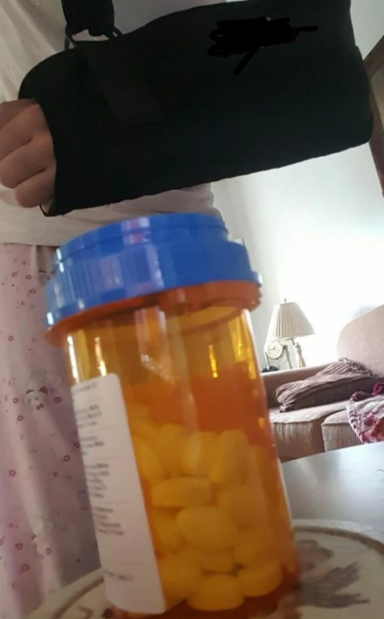 Osoba s rukou v ortéze vedľa fľaštičky s liekmi v obývacej izbe so svetlom lampy v pozadí.