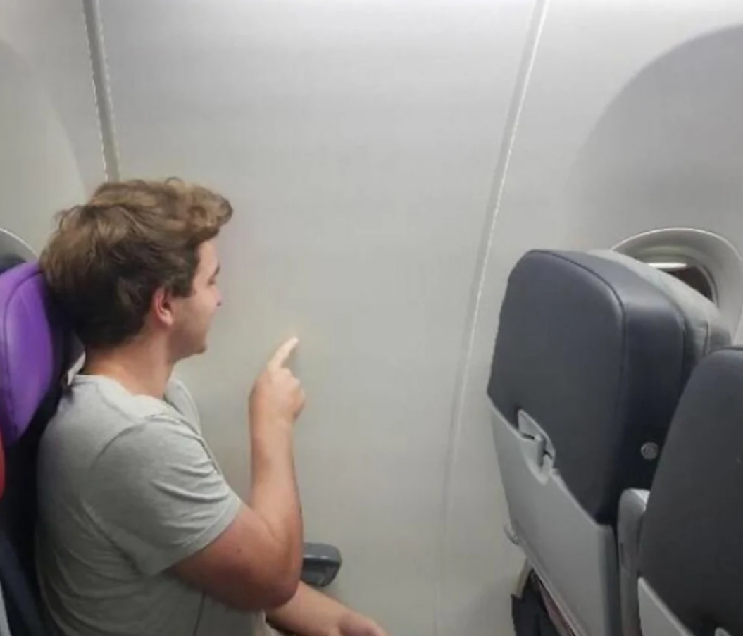 Cestujúci sedí v lietadle bez okna, ukazuje na stenu a usmieva sa vtipnej situácii.