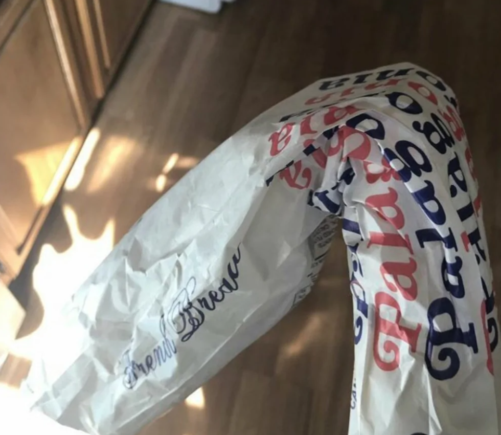 Ohýbaný papierový obal na bagetu s textom „French Bread“ v kuchyni, podsvietené slnečnými lúčmi.