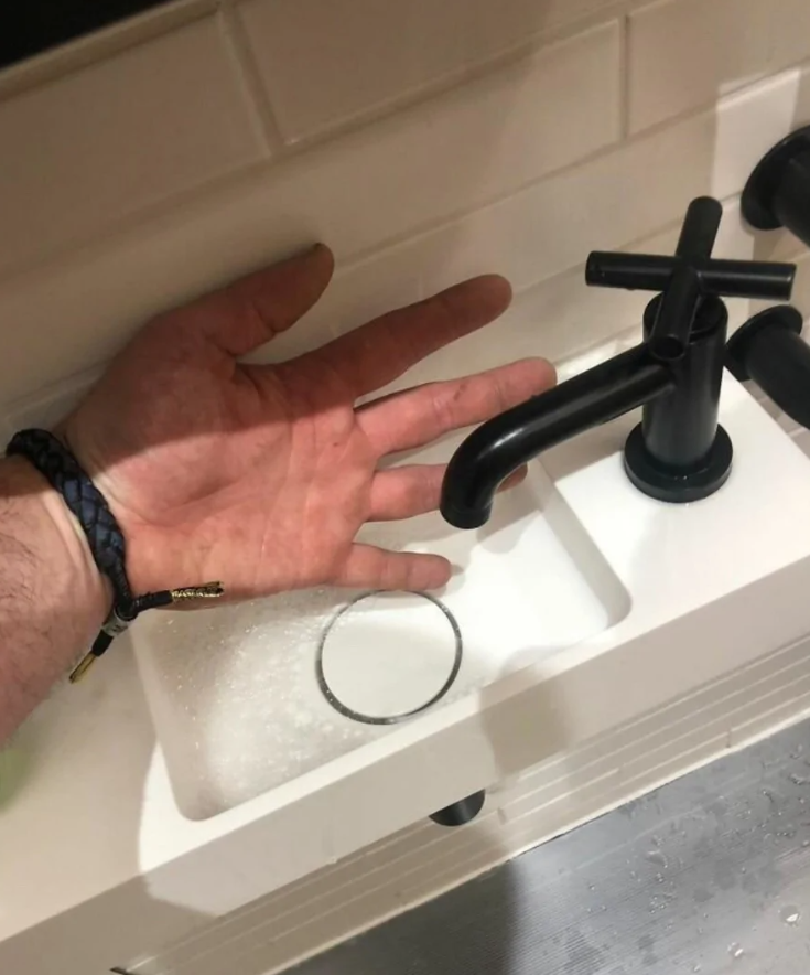 Ruka pod malým čiernym kohútikom nad kompaktným bielym umývadlom s bielym obkladom v pozadí.