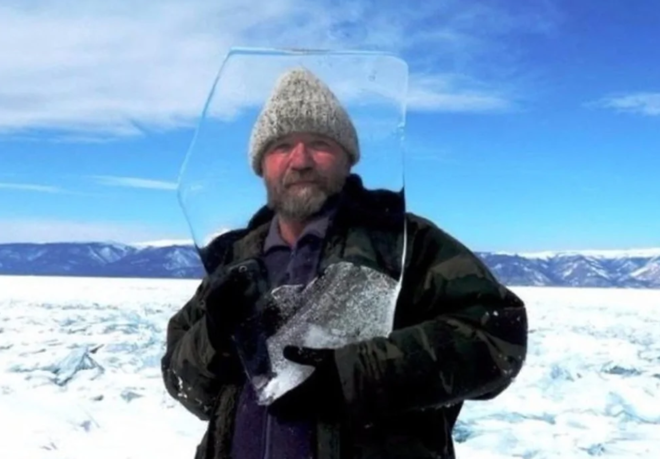 Muž v zimnom oblečení drží kus ľadu na zamrznutom jazere s horami v pozadí.