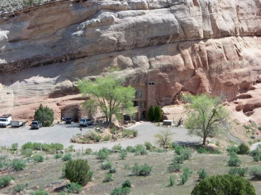 Dom včlenený do útesu s parkoviskom a stromami, zasadený do púštnej krajiny.