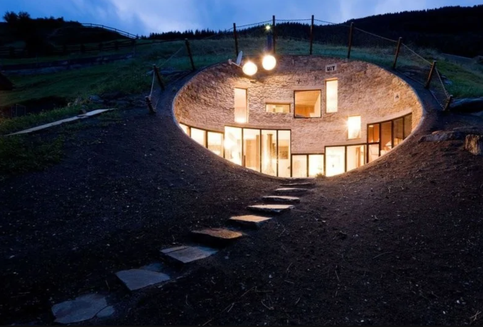 Podzemný dom so svietiacimi oknami integrovaný do svahu, schody vedú k jeho modernému vchodu v noci.