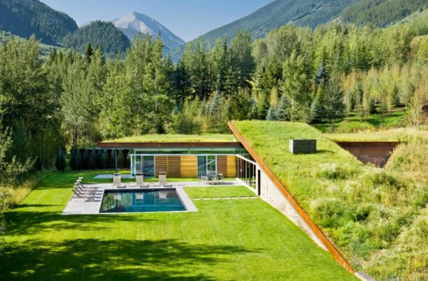 Moderný dom s trávnatou strechou a bazénom v lese, obklopený horami a zelenou prírodou.