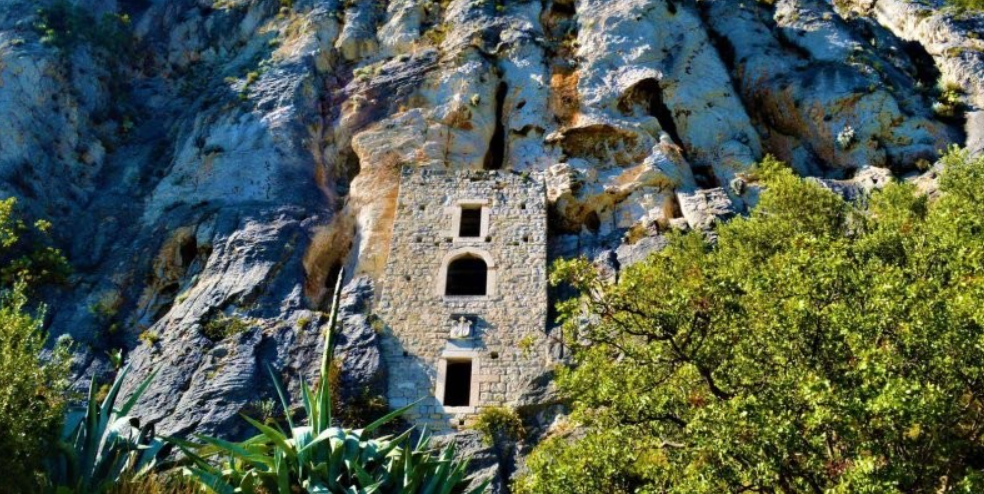 Kamenná budova včlenená do skaly, obklopená zeleňou a stromami, na slnečnom pozadí skalného útesu.