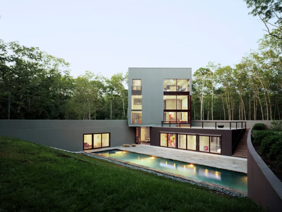 Moderný dom s bazénom a veľkými oknami obklopený zeleňou, minimalistická architektúra v prírodnom prostredí.