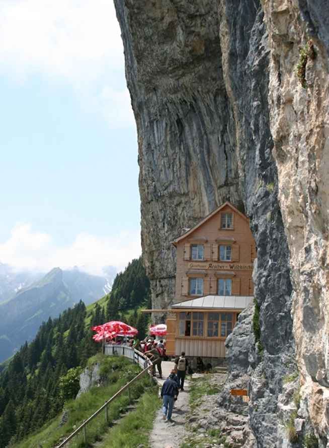 Horský hostinec prilepený na úbočí skaly, s turistami na horskom chodníku a výhľadom na zelené lesy a hory.