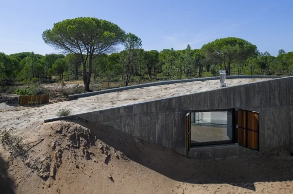 Moderný domček v piesočnatom teréne obklopený zeleným lesom.