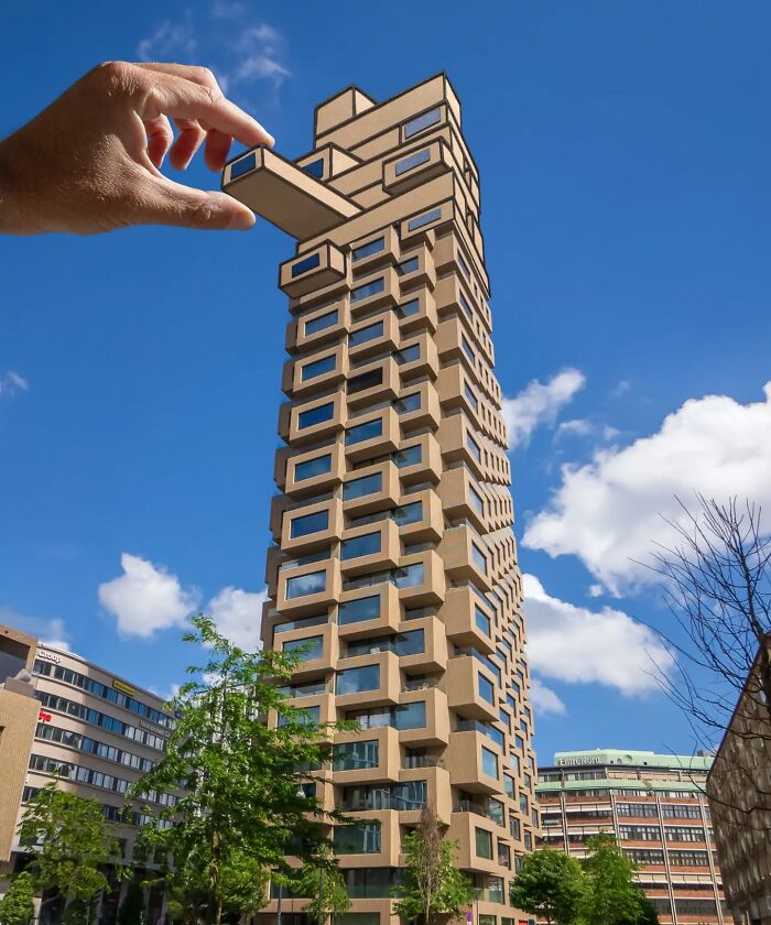 Ruka hrajúca Jenga s modernou budovou, optická ilúzia na modrom nebi.