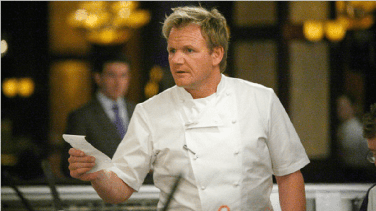 Odborný šéfkuchár v bielej uniforme drží papier a sústredene komunikuje v reštauračnom prostredí.