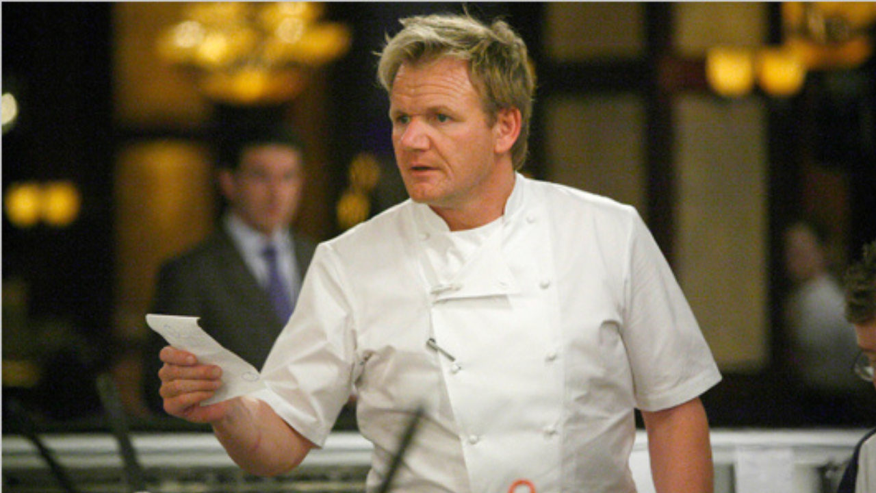 Odborný šéfkuchár v bielej uniforme drží papier a sústredene komunikuje v reštauračnom prostredí.