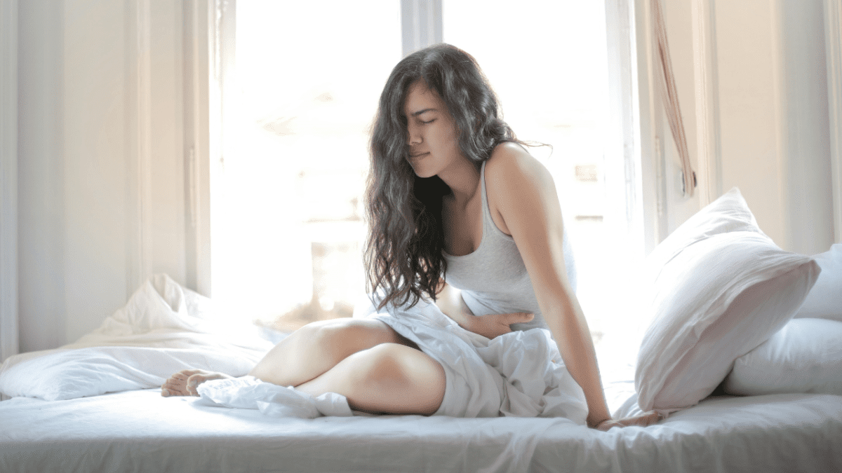 Žena s bolesťami brucha sedí na posteli pri dennom svetle.