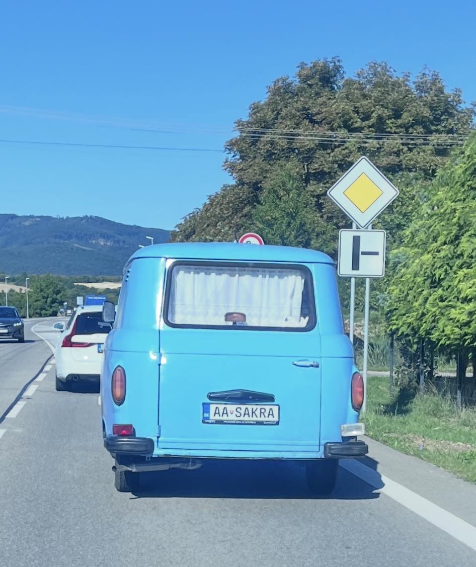 Modrá dodávka s vtipnou poznávacou značkou na ceste v krásnom slnečnom dni, obklopená stromami a značkami.