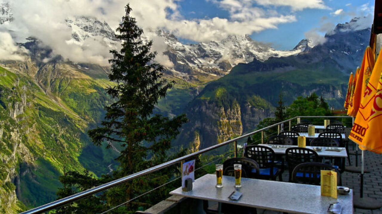 Horská terasa s výhľadom na zasnežené vrcholy, prázdne stoly a žlté slnečníky v alpskom prostredí.