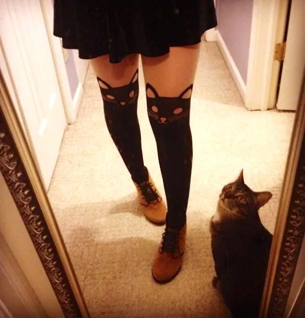 Osoba v pančuchách s mačacím vzorom stojí pred zrkadlom, vedľa nej sedí sivá mačka.