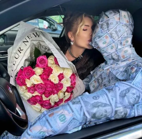 Žena bozkávajúca osobu zabalenú v bankovkách v aute, držiac kyticu ruží.