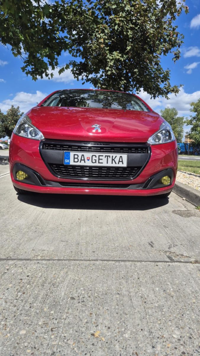 Červené auto Peugeot zaparkované pod stromom na slnečnej ulici.