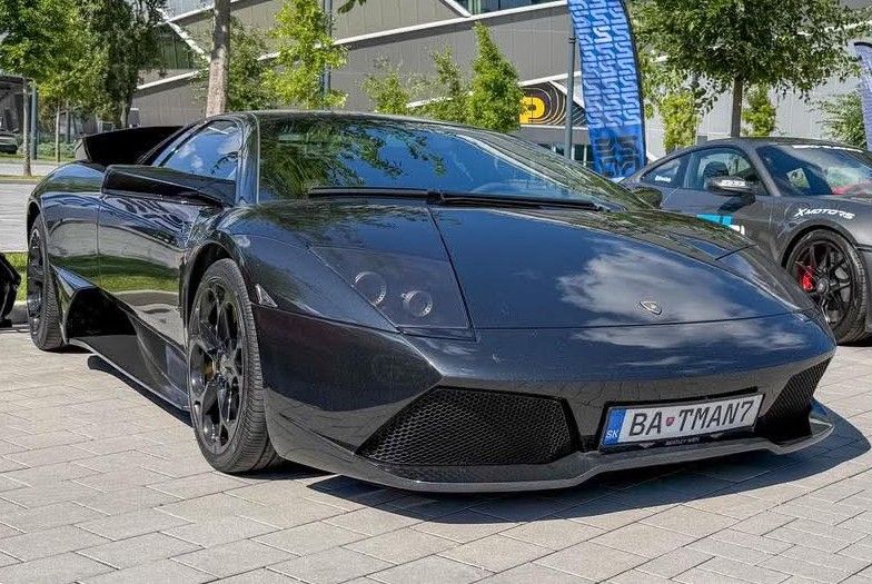 Čierne športové auto zaparkované na ulici pod slnečným nebom, s viditeľnou ŠPZ a ďalšími autami v pozadí.