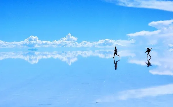 Dvaja ľudia kráčajú po zrkadlovo lesklom povrchu slaných plání pod modrou oblohou s oblakmi.