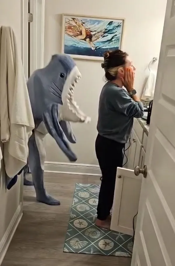 Osoba v kúpeľni prekvapená kostýmom žraloka vtipne sa plížiacim zozadu.