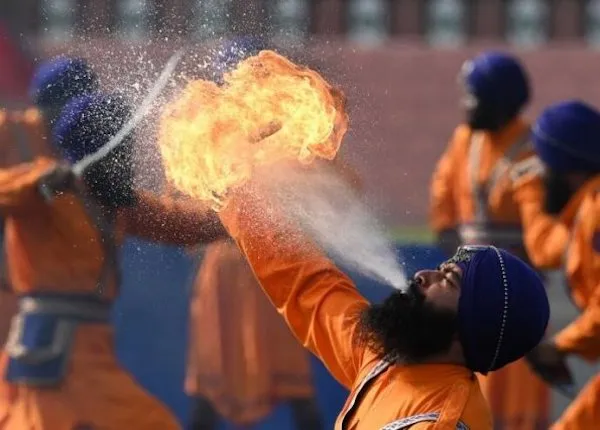 Muž v oranžovom tradičnom oblečení predvádza ohňovú show, vydychujúc plamene počas kultúrneho podujatia.