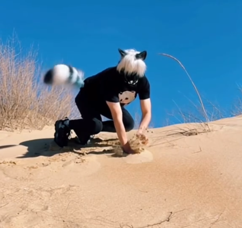 Osoba v maske zvieraťa hrá sa v piesku na slnečnom kopci, jasná obloha a suché rastliny na pozadí.