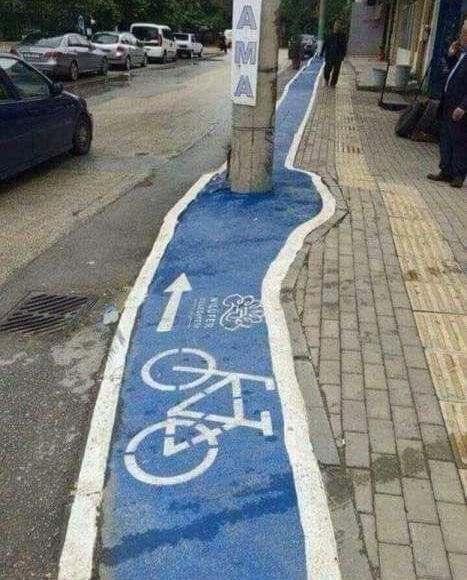 Krivolaká cyklotrasa na chodníku s prekážkami, obklopená autami, stĺpmi a budovami v mestskom prostredí.