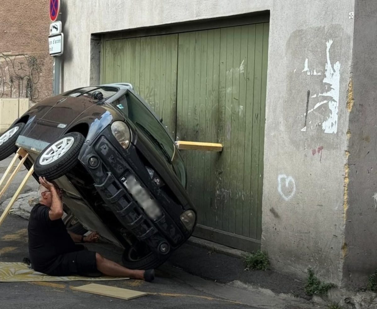 Auto opreté o garážové dvere, muž leží vedľa neho na zemi, zdanlivo kontroluje situáciu.