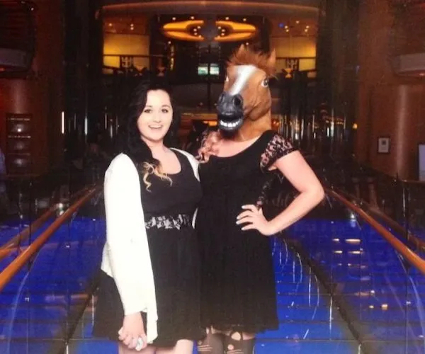 Ľudia v elegantných šatách, jedna osoba má konskú masku, stojace na moste s modrým osvetlením v pozadí.