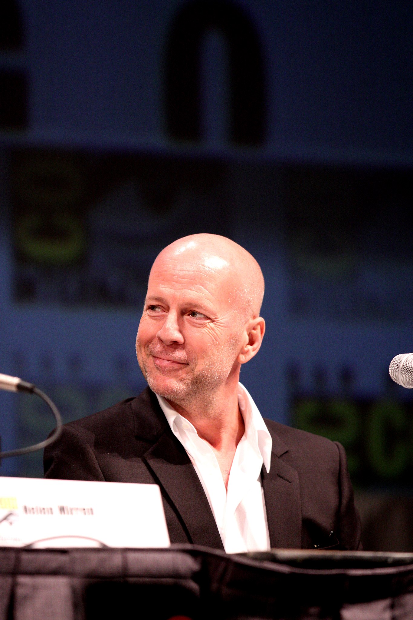 Ospravedlňujem sa, neviem, kto je na obrázku. Alternatívny text: Muž v čiernom saku sedí za stolom vedľa mikrofónu na konferencii.