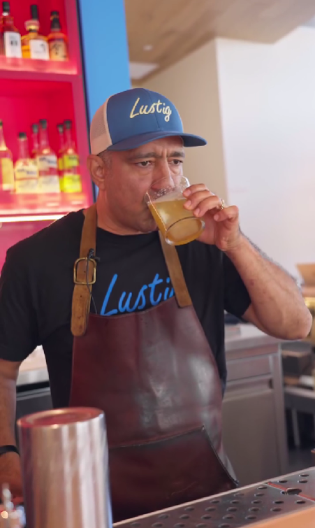 Muž v čiapke a zástere ochutnáva nápoj za barovým pultom, v pozadí regál s alkoholom.