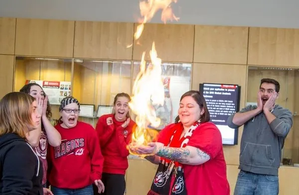 Skupina ľudí prekvapene sleduje veľký plameň vychádzajúci z dlane ženy v červenom oblečení.